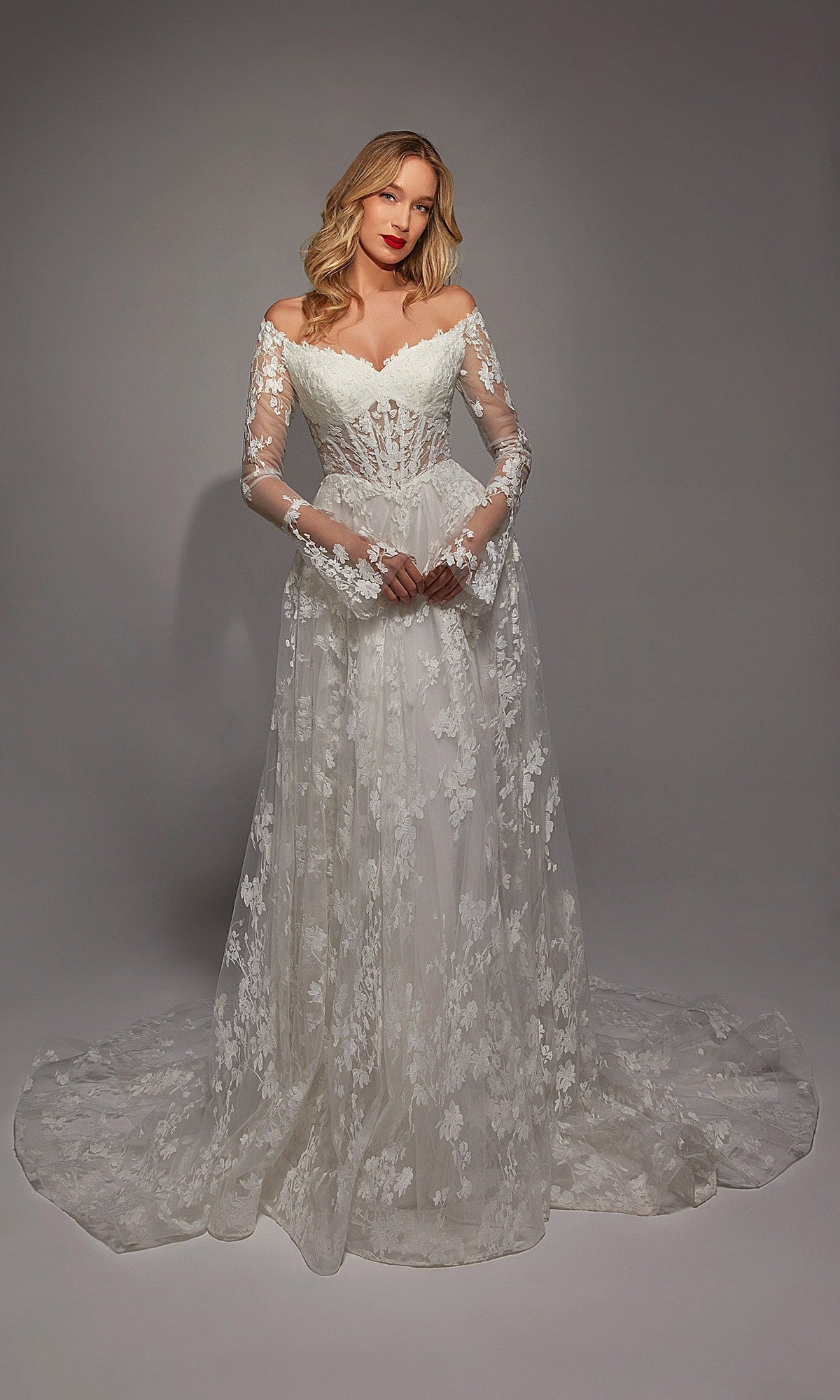 A woman poses in a studio wearing the Alyce 7150, a long white dress featuring off-the-shoulder long sleeves, white lace with ruffle details on the fitted bodice, and a flowing train.